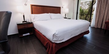 Habitación de hotel con una cama grande, cabecera de madera, dos mesitas de noche con lámparas, una silla y vistas a zonas verdes por la ventana.