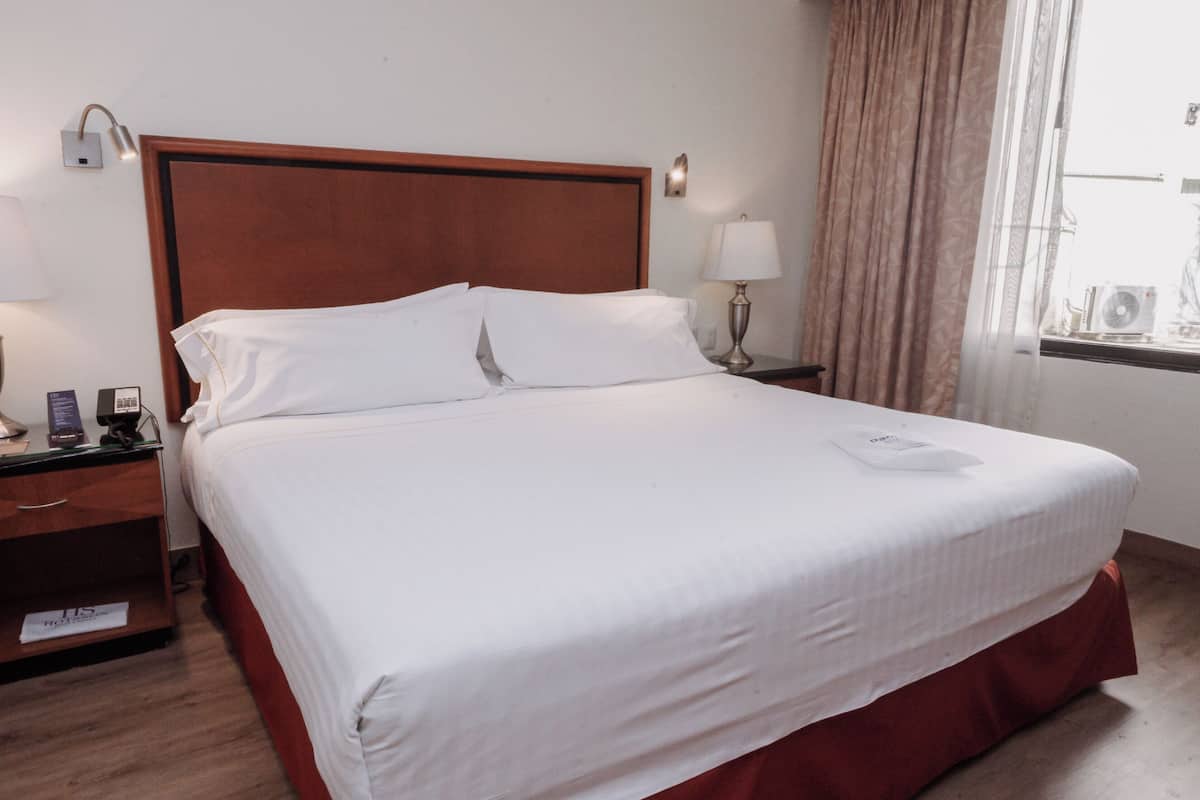 A hotel room with a wooden headboard, two bedside tables with lamps, a bed with white linens, and a window with curtains.