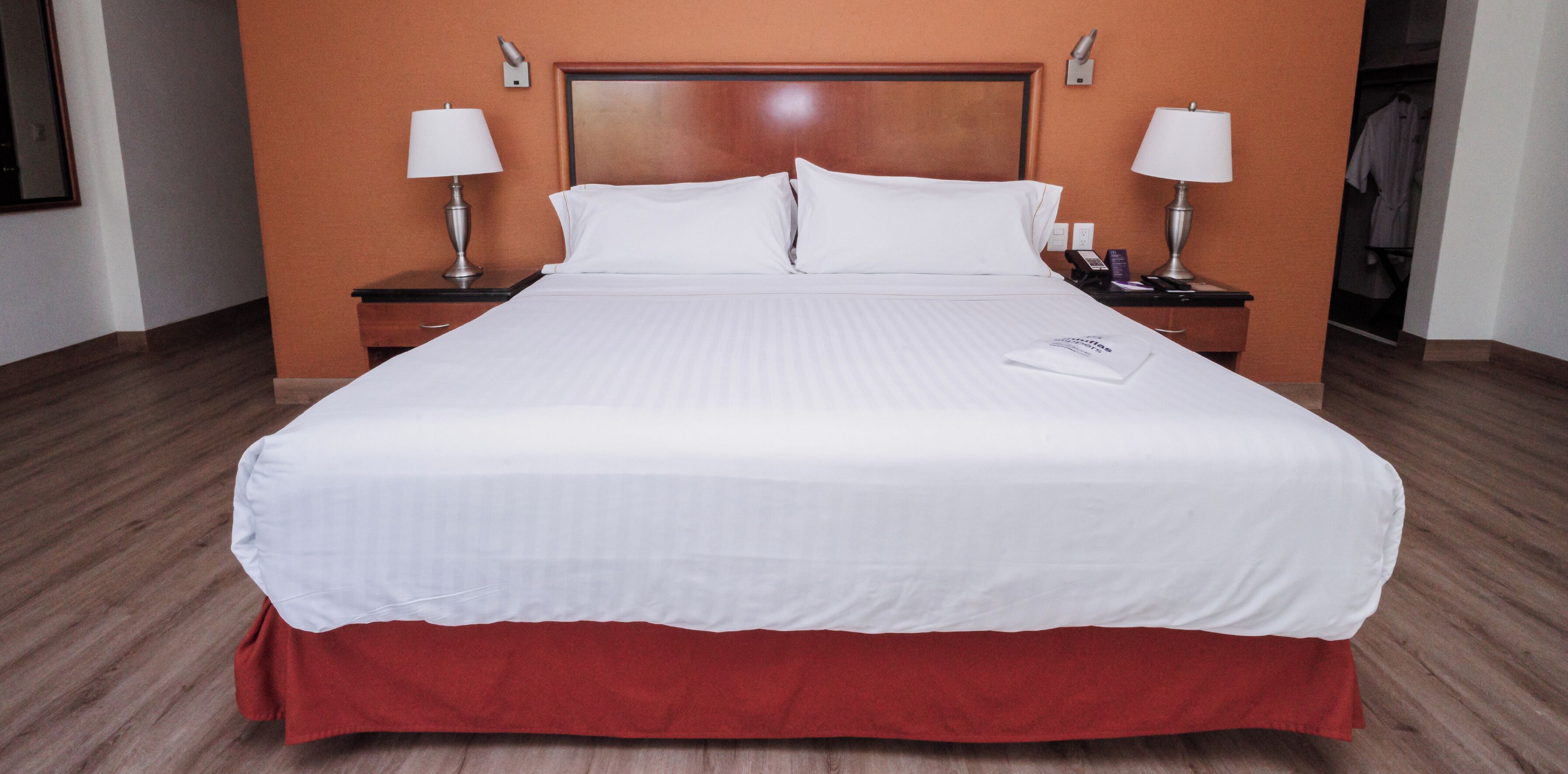 Habitación de hotel con una cama grande, cabecera de madera, dos mesitas de noche con lámparas y una colcha roja que cubre el borde de la cama.