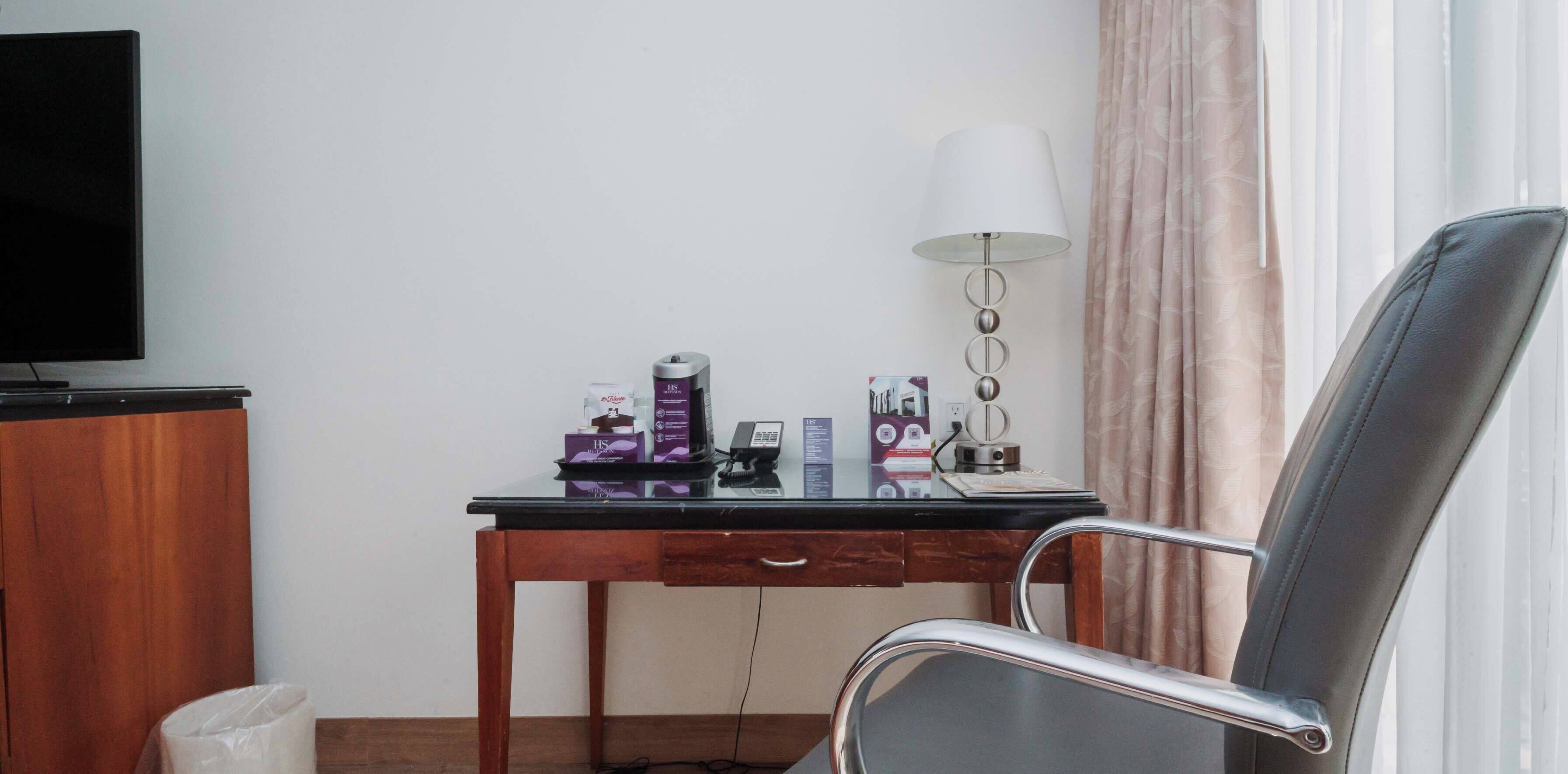 A hotel room with a wooden desk, a black chair, a TV, a lamp, and a window with curtains.