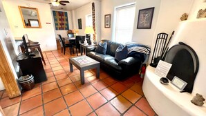 Un salon comprenant un canapé, une table basse et une cheminée. On y trouve également un coin repas avec une table et des chaises. Le sol est carrelé et la pièce est équipée d’un ventilateur de plafond.