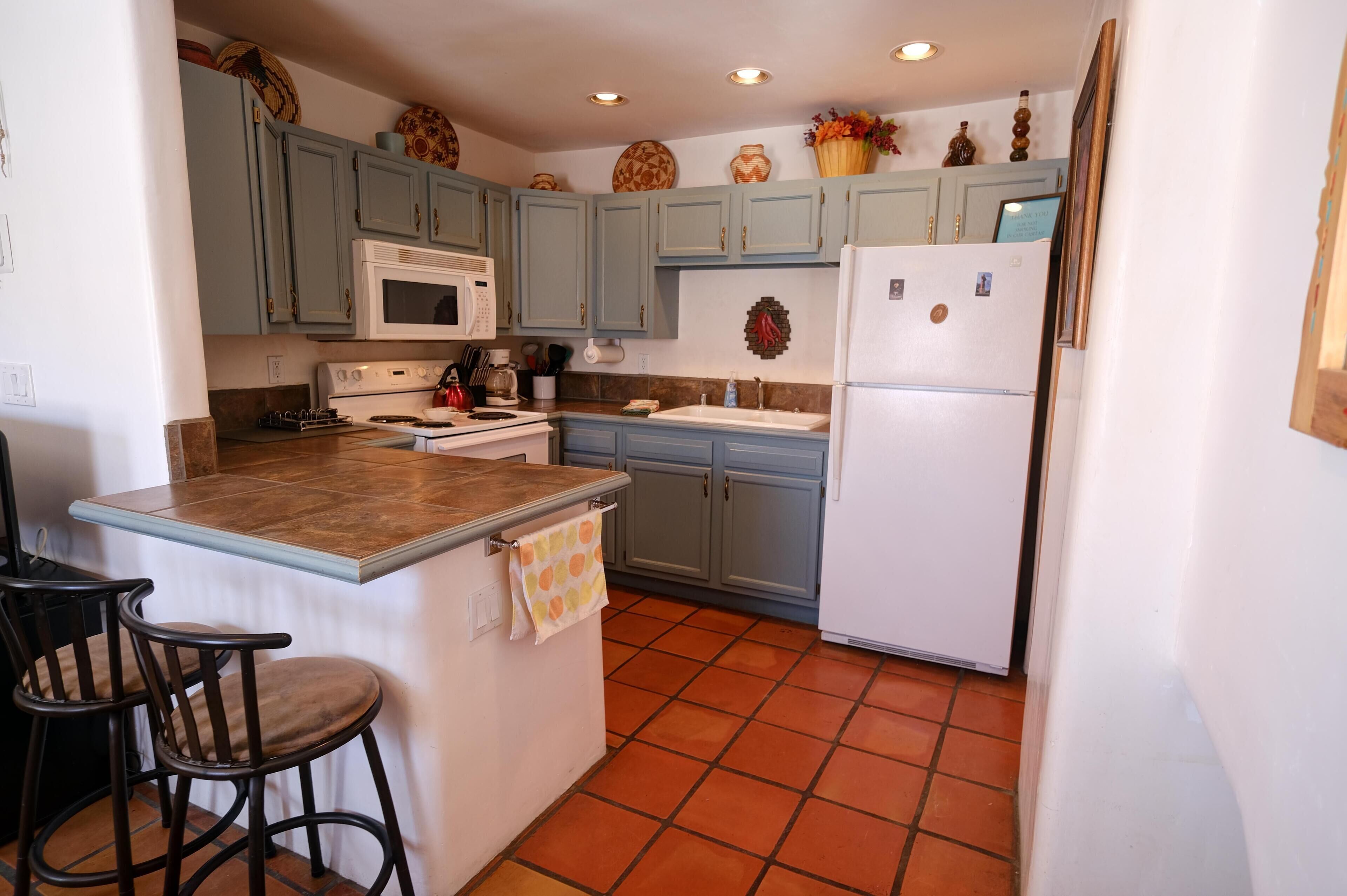 A kitchen with light blue cabinets, a white refrigerator, a microwave, and a stove. There are two bar stools and a small island with a towel.