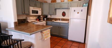 A kitchen with light blue cabinets, a white refrigerator, a microwave, and a stove. There are two bar stools and a small island with a towel.
