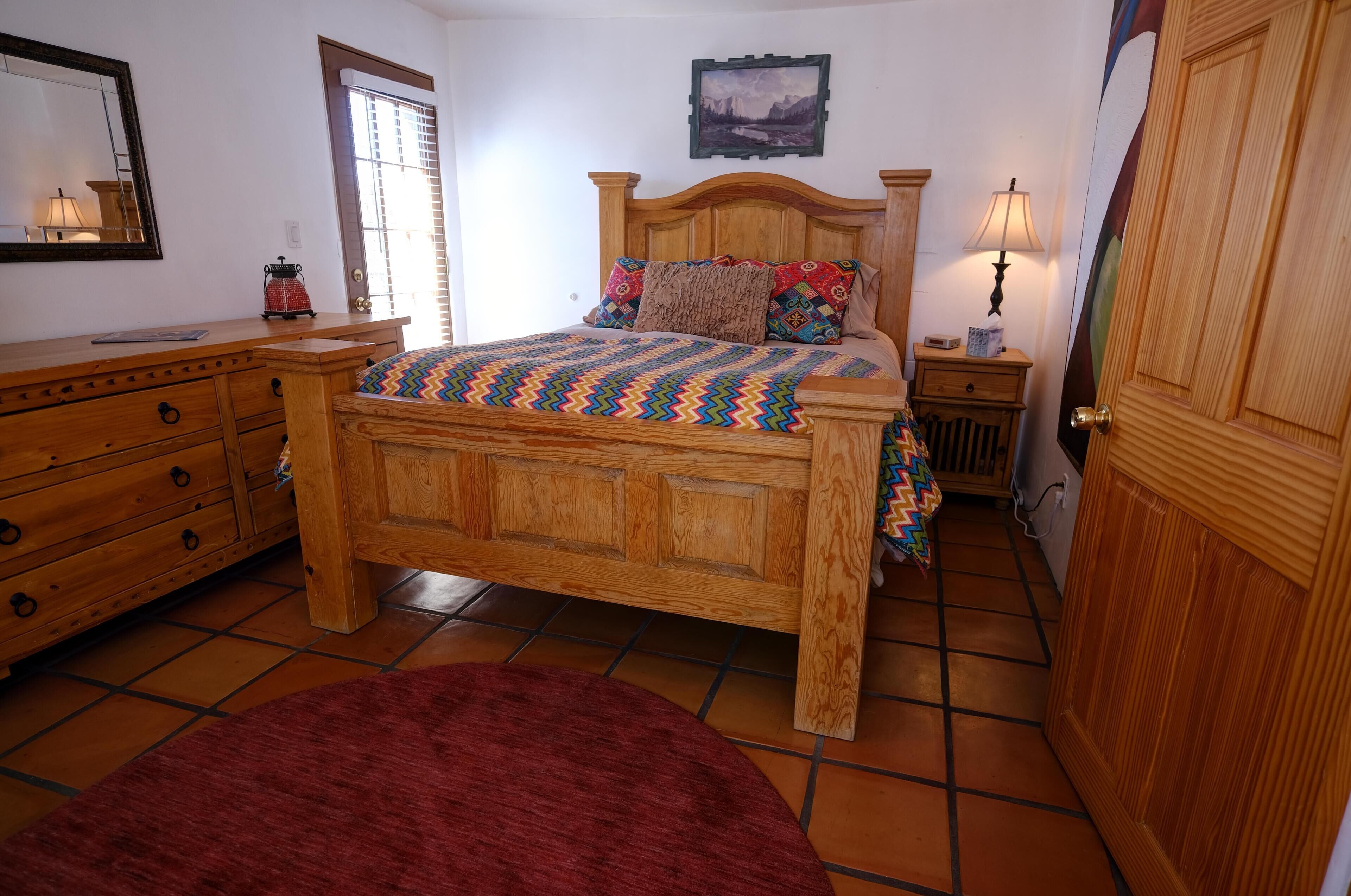 Een slaapkamer met een houten bed, een dressoir, een rood vloerkleed, een nachtkastje met een lamp en een deur met een houten paneel.
