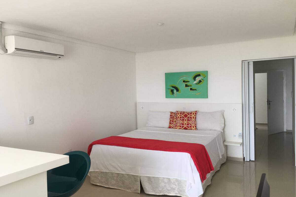 A hotel room with a bed, a desk, and a chair. There is a painting on the wall and an air conditioning unit mounted on the wall.