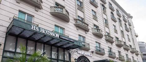 La façade d’un hôtel présente un style architectural classique, avec de nombreux balcons et une enseigne bien visible indiquant « HS HOTSSON ».