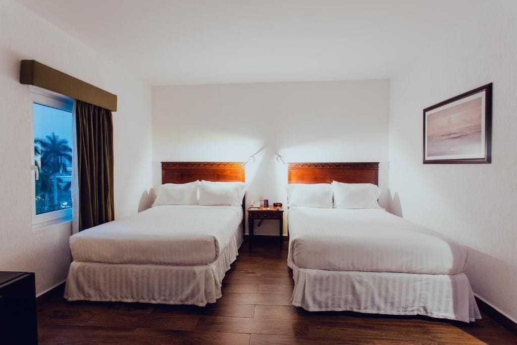Una habitación de hotel con dos camas, una mesita de noche, una ventana con cortinas y un cuadro en la pared.