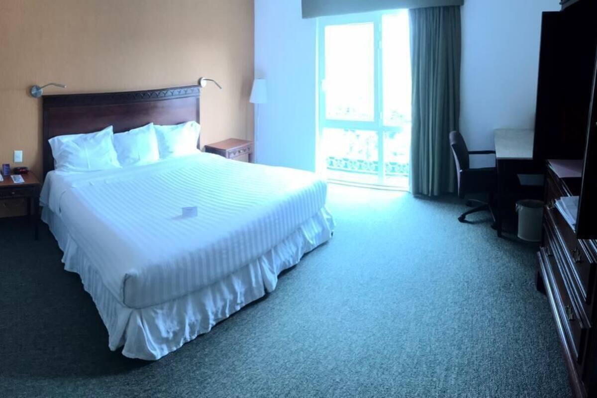Habitación de hotel con una cama grande, un escritorio, una silla, un televisor y una ventana con cortinas.
