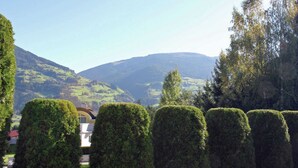 On aperçoit des haies soigneusement taillées et un bâtiment avec un toit rouge, entouré de collines verdoyantes et de montagnes en arrière-plan.