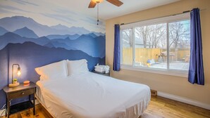A bedroom with a bed, a ceiling fan, a nightstand, a lamp, a window with curtains, and a mountain mural on the wall. - Stir Crazy! Bike Trails, Dogs OK, Food & Nightlife (Colorado Springs)
