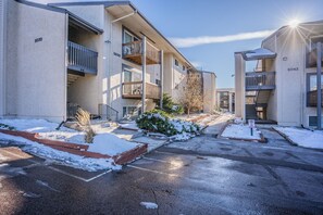 A multi-story residential building with numbered units, a snow-covered driveway, and a clear sky.