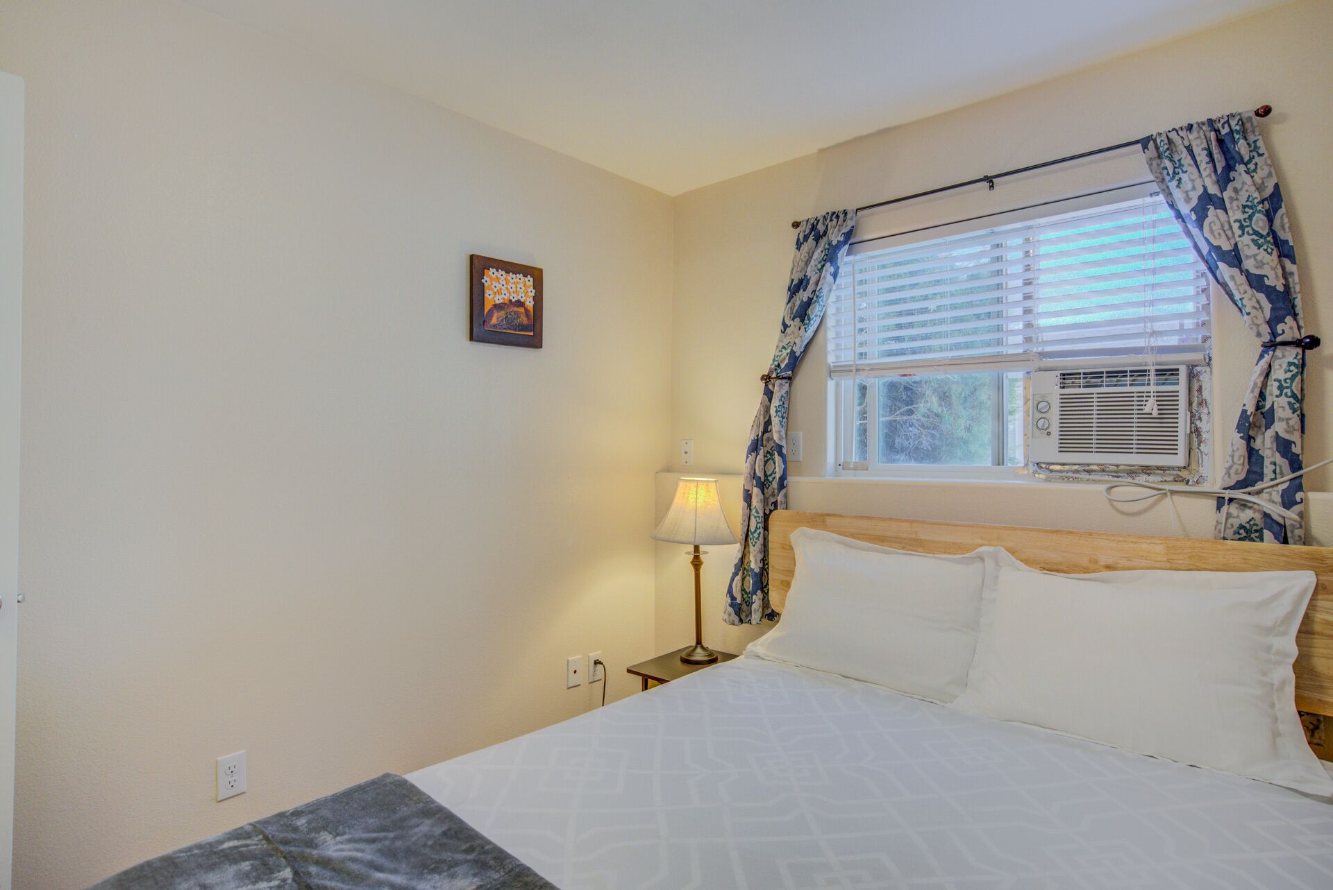 Un dormitorio con cama, una mesita de noche, lámpara, ventana con cortinas, equipo de aire acondicionado y un cuadro colgado en la pared.