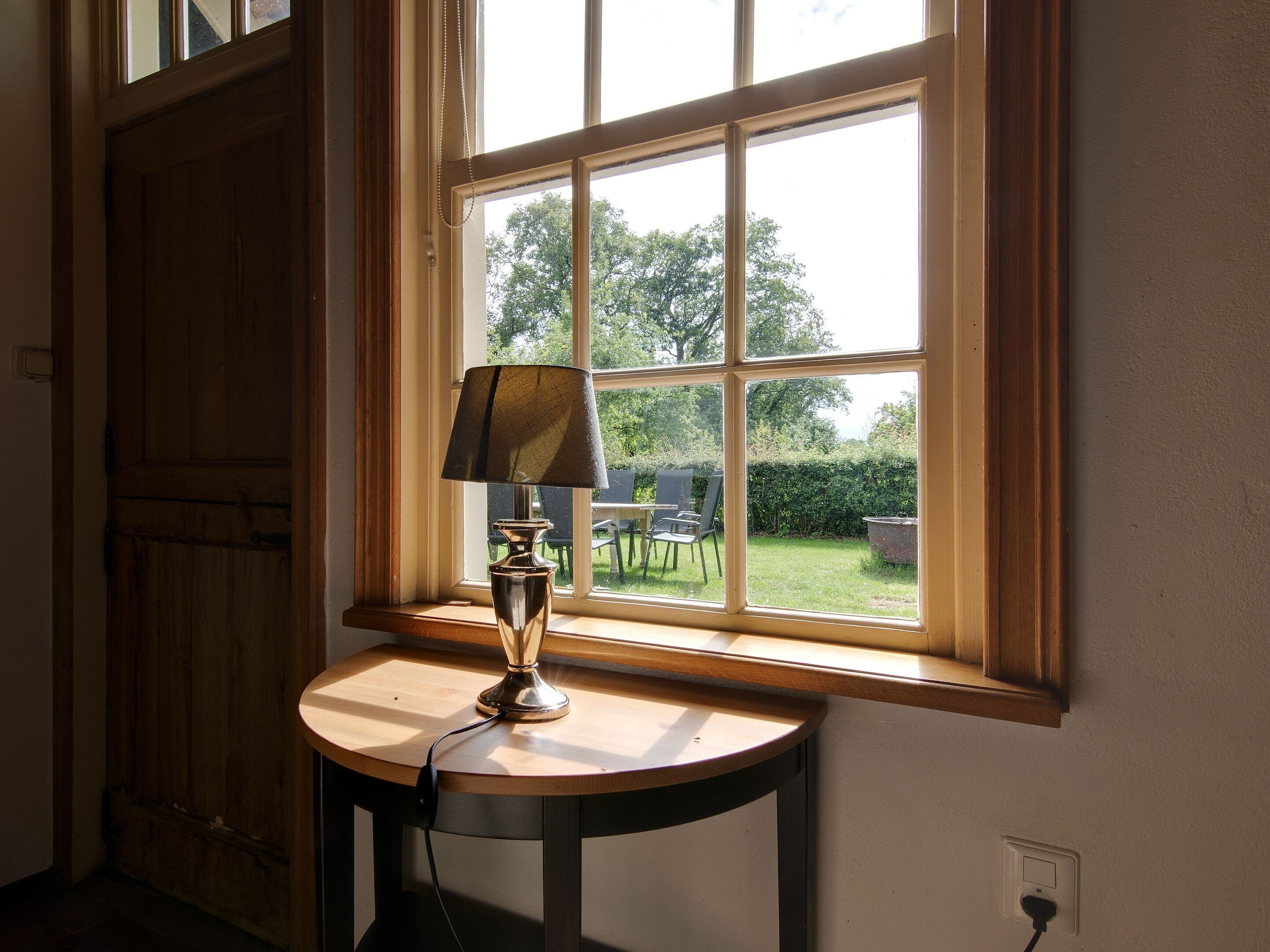 Una lámpara sobre una mesa junto a una ventana que ofrece vistas a un jardín y mobiliario exterior.