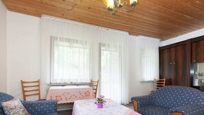 Une pièce chaleureuse avec un plafond en bois, deux canapés bleus à motifs, une petite table recouverte d’une nappe rose et une fenêtre ornée de rideaux blancs.