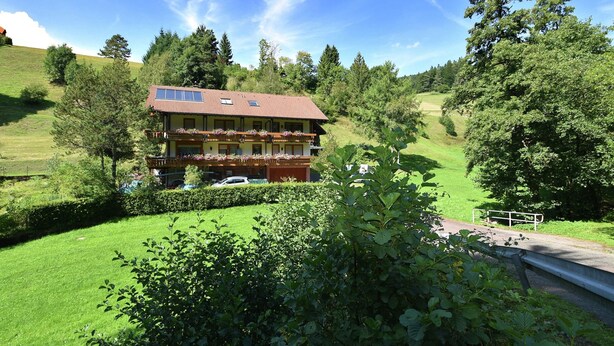 Un bâtiment à deux étages, doté d’un toit recouvert de tuiles rouges, de balcons ornés de jardinières et offrant une vue sur un paysage verdoyant, avec des arbres et une colline en arrière-plan.