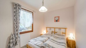 A bedroom with a wooden bed, a window with patterned curtains, a bedside table with a lamp, and a wall-mounted picture. - Gartenbungalow in Blankenfelde-mahlow (Blankenfelde-Mahlow)