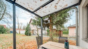 A covered patio with a table and chairs, a metal railing, and a view of a backyard with trees and a house. - Gartenbungalow in Blankenfelde-mahlow (Blankenfelde-Mahlow)