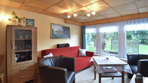 Uma sala de estar aconchegante com um sofá vermelho, um armário de madeira, uma mesa de jantar com toalha rendada e janelas amplas com vista para o jardim.