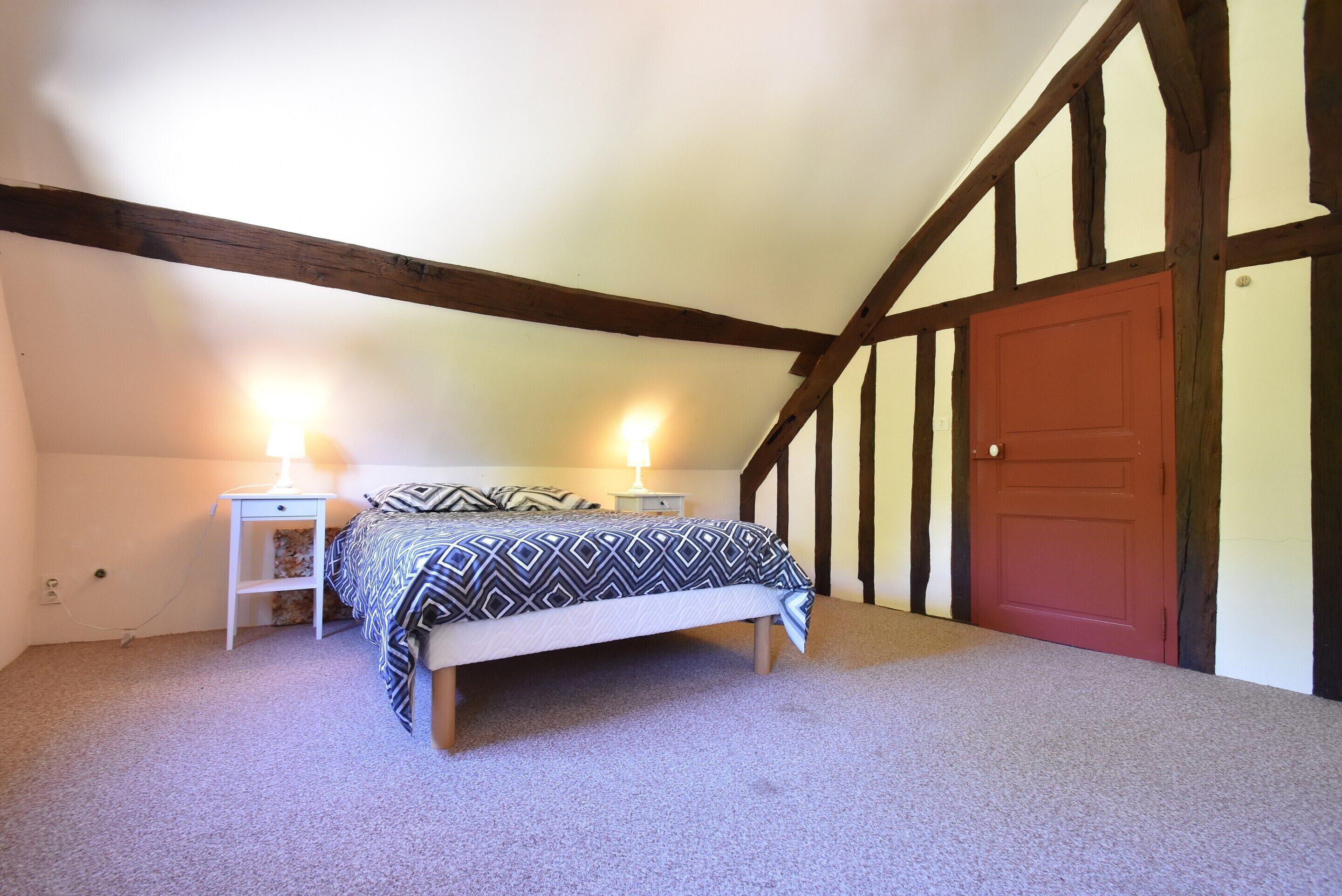 Une chambre à coucher avec un lit, deux tables de chevet, une commode, une porte rouge et des poutres apparentes.