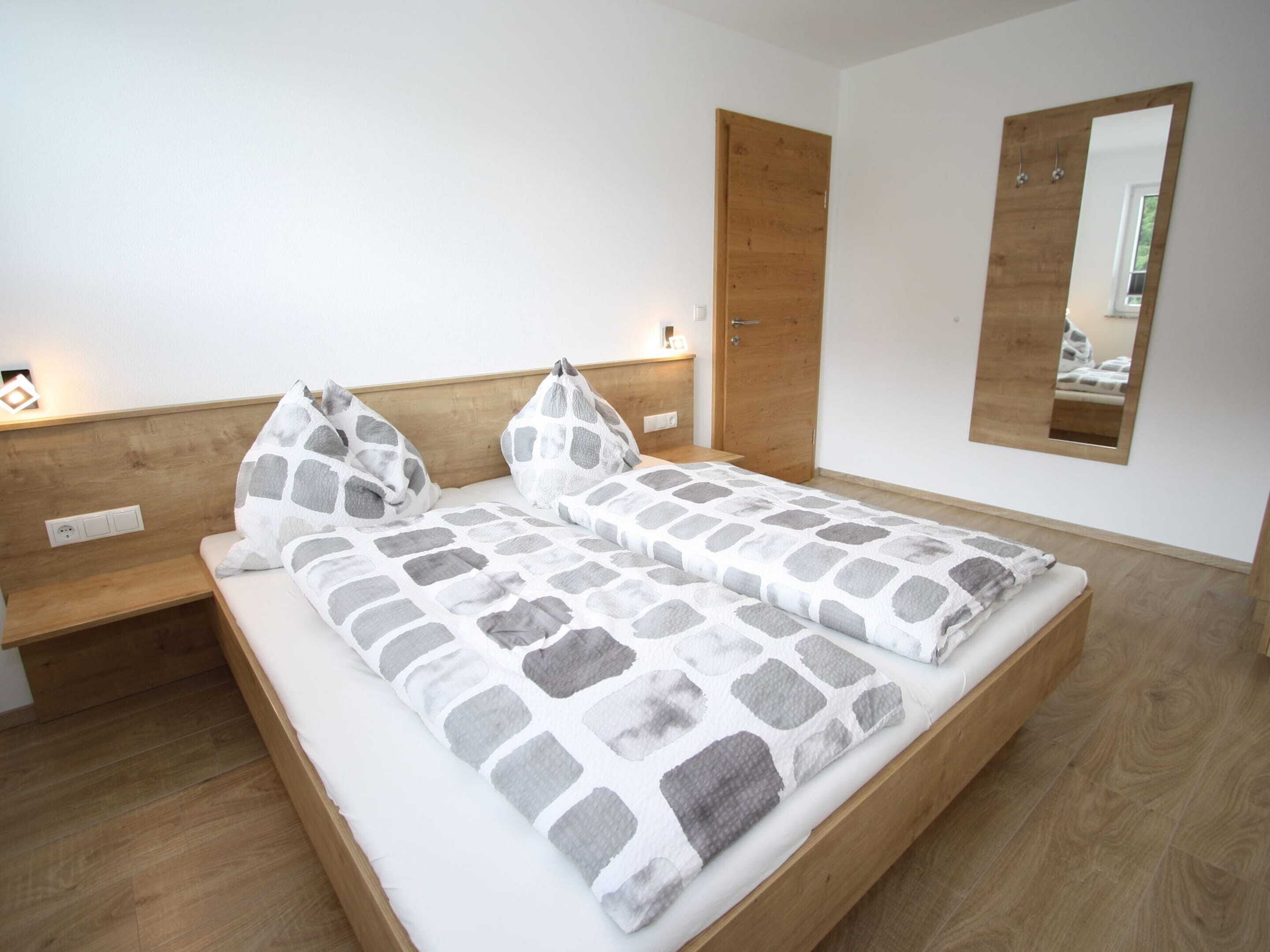 Un dormitorio con cama de madera, dos almohadas, cabecero de madera, mesita de noche, espejo y enchufe en la pared.