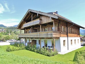 Une maison traditionnelle en bois, dotée d’un balcon, entourée d’une pelouse bien entretenue et avec une chaîne de montagnes en toile de fond.