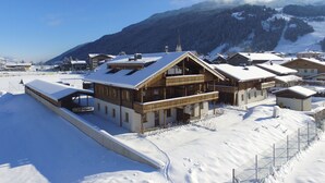 A wooden chalet with a balcony, surrounded by snow-covered buildings and mountains in the background. - Apartment in Hollersbach With Sauna Near ski Area (Hollersbach im Pinzgau)