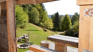 A wooden deck with picnic tables and a fire pit, overlooking a grassy hillside with trees and a clear sky.