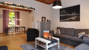 Ein modernes Wohnzimmer mit grauem Sofa, weißem Couchtisch und schwarzem Sessel. Ein Essbereich mit Tisch und Stühlen ist vorhanden, und im Hintergrund ist eine Küche erkennbar.