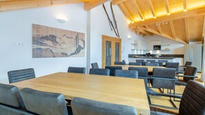 Et moderne spiseområde med et stort træbord og grå polstrede stole, et køkken i baggrunden og et maleri af et bjerglandskab på væggen.