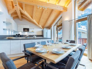 Una cocina moderna con vigas de madera, una zona de comedor con una mesa grande dispuesta para seis personas, y vistas a montañas a través de ventanas amplias.