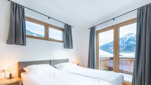 Et soveværelse med to senge, et trænatbord, en sengetavlelampe og udsigt til sneklædte bjerge gennem store vinduer.