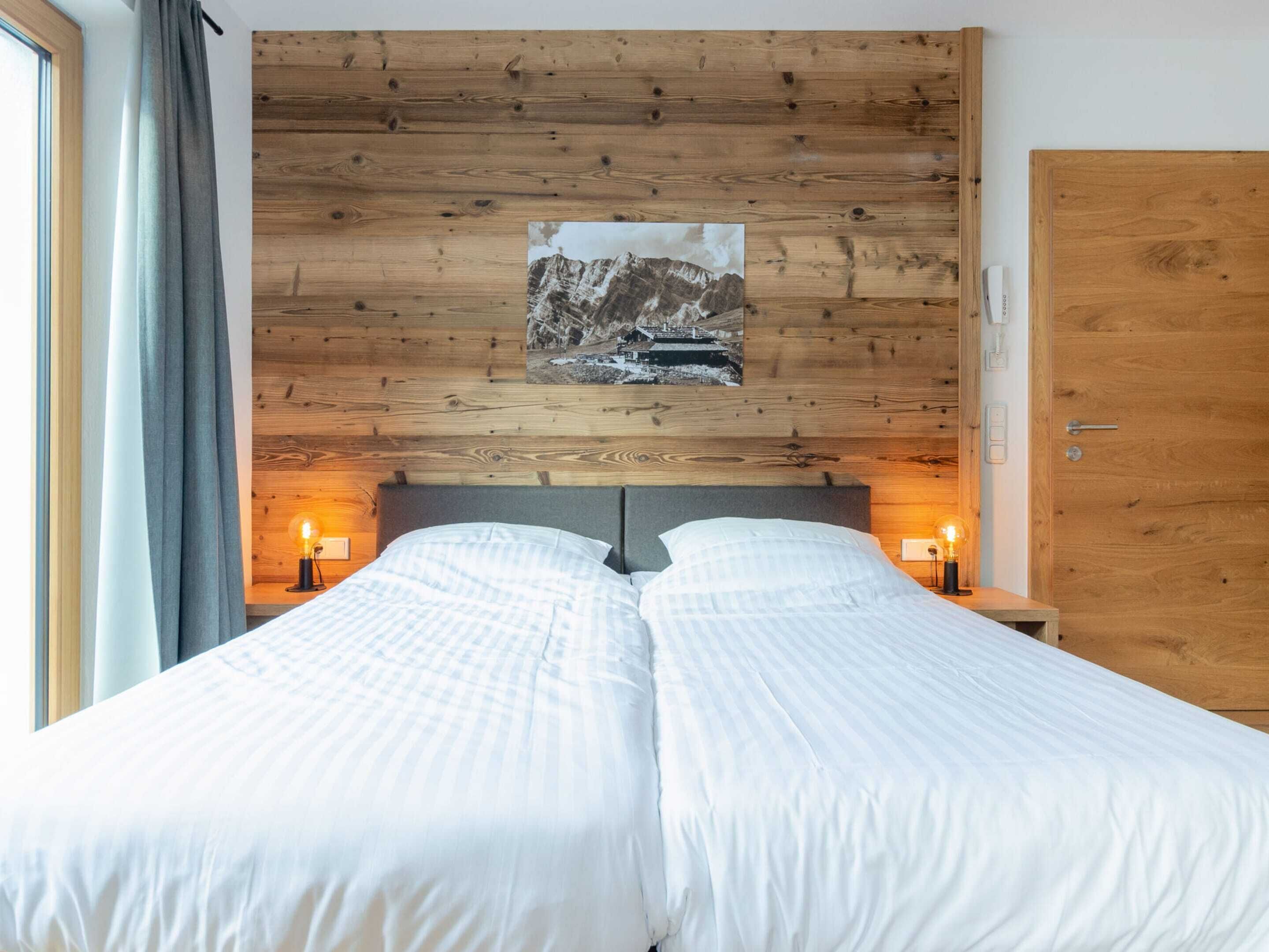 A double bed with white bedding, a wooden headboard, two bedside lamps, and a framed picture of a mountain landscape.