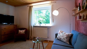 A cozy living room with a wooden ceiling, a television, a sofa, a chair, a small table with a vase, and a window with a view of greenery. - Apartment With Sauna (Zandt)