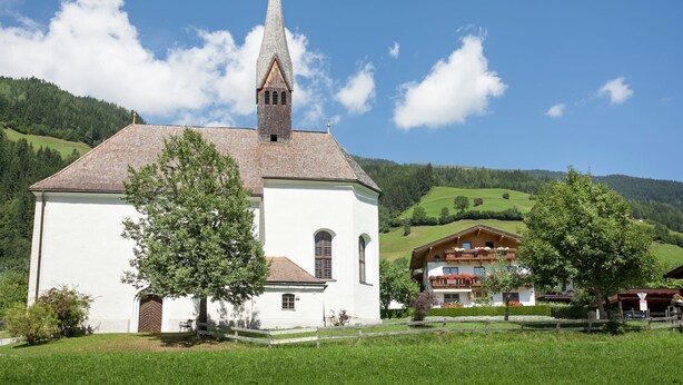 A white church with a tall steeple and a bell tower, surrounded by green hills and a clear blue sky. - Beautiful Apartment With Large, Sunny Balcony (Hollersbach im Pinzgau)