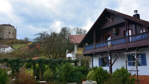 A traditional wooden house with a balcony, surrounded by lush greenery and a historic building in the background. - Ferienhaus in Saldenburg mit Sauna (Saldenburg)