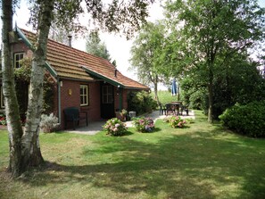 Een bakstenen gebouw met een groen dak, een terras met buitenmeubilair en een verzorgde tuin met bloemen en struiken.