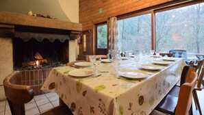 A dining area with a long table set for four, a fireplace, and a view of trees through large windows. - Family Villa With Garden (Bouillon)