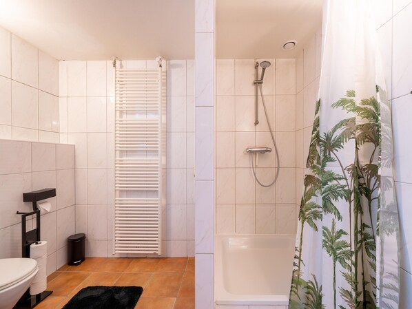 Un baño moderno con inodoro, un cubo de basura negro, un toallero blanco, un radiador blanco, una ducha con cabezal de ducha manual y una alfombra negra.