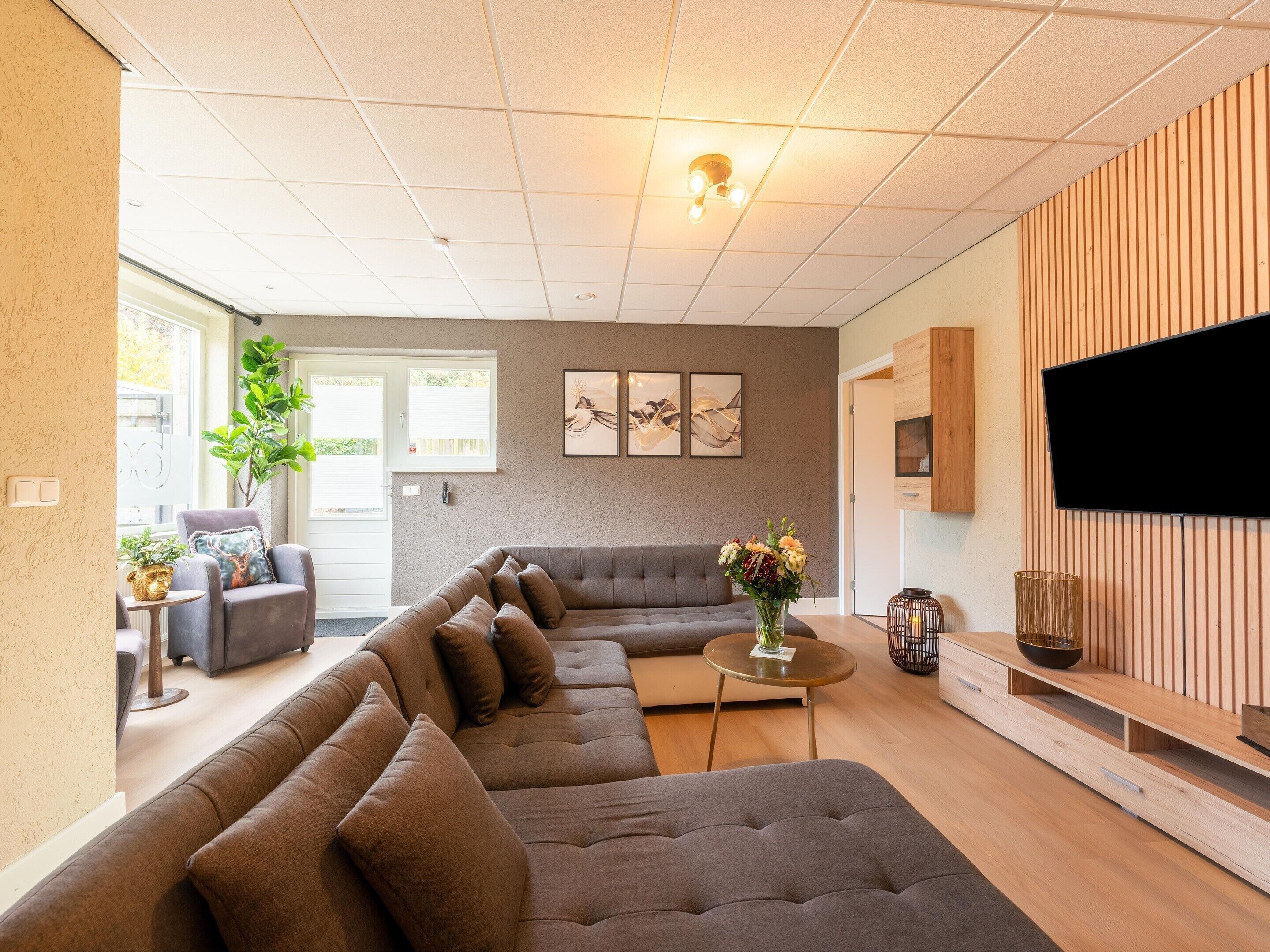 A modern living room with a sectional sofa, a flat-screen TV mounted on a wooden wall, and a small round table with a vase of flowers.