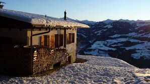 A snow-covered mountain house with a stone wall and wooden shutters, set against a backdrop of snow-covered mountains and a clear sky.