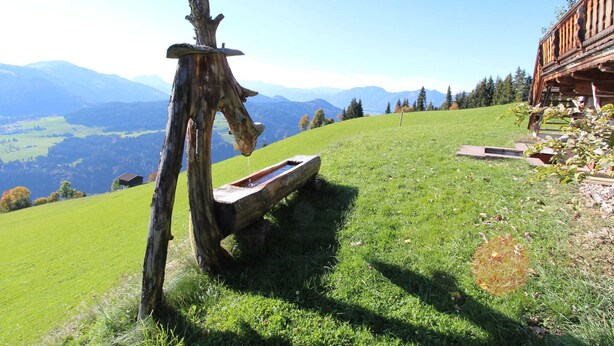 A rustic wooden bench on a grassy hillside with a wooden structure in the background and a mountainous landscape in the distance.