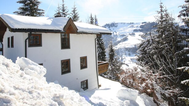 A white chalet with multiple windows and a snow-covered roof, surrounded by snow-laden trees and a mountainous backdrop. - Boutique Chalet in Maria Alm near Hochkönig (Maria Alm am Steinernen Meer)