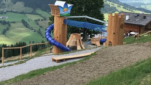 A playground with a slide, climbing net, and wooden structures, set against a backdrop of mountains and a building. - Boutique Chalet in Maria Alm near Hochkönig (Maria Alm am Steinernen Meer)