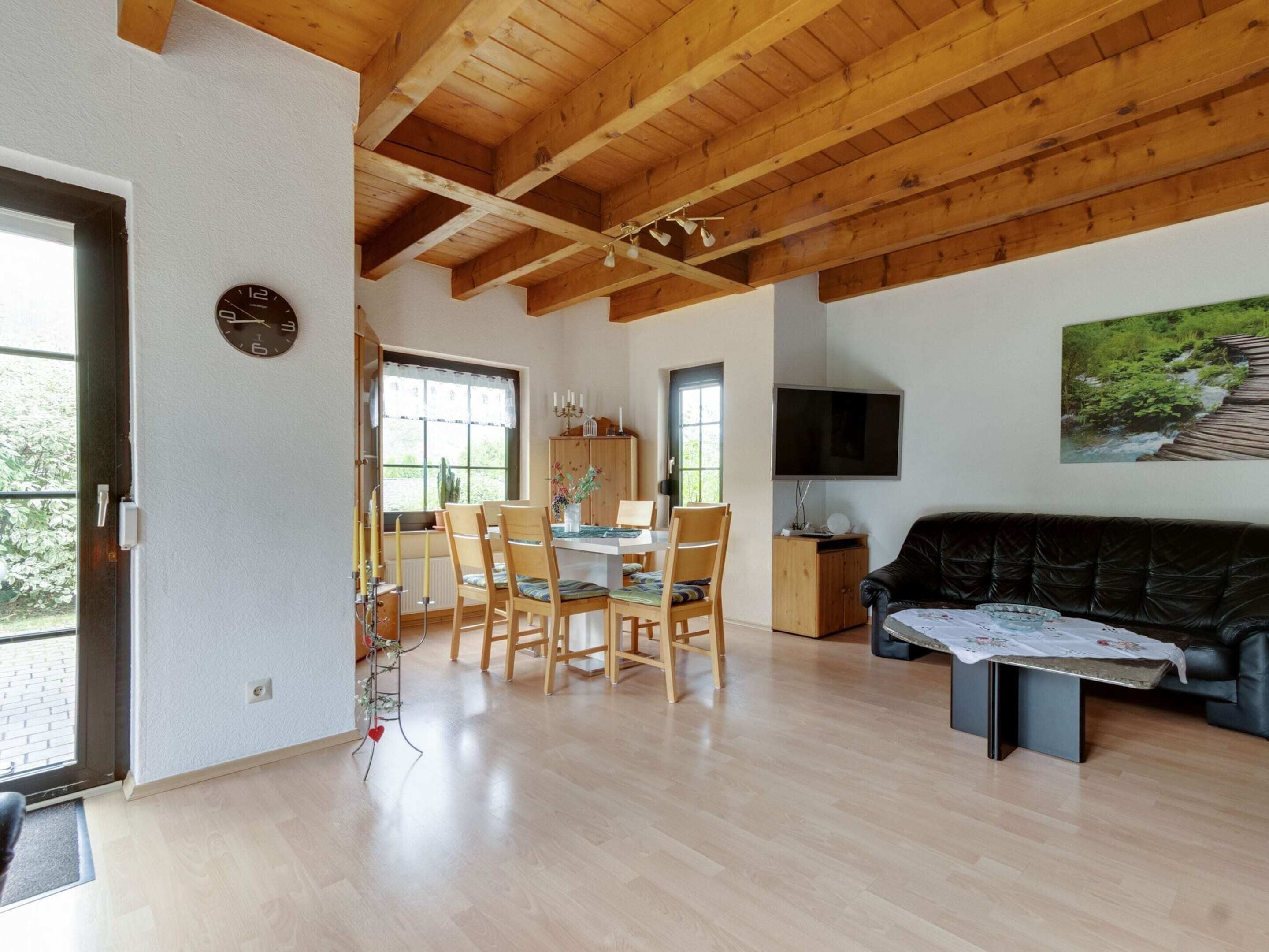 A spacious living area with a wooden ceiling, a dining table with chairs, a black leather sofa, and a flat-screen TV mounted on the wall.