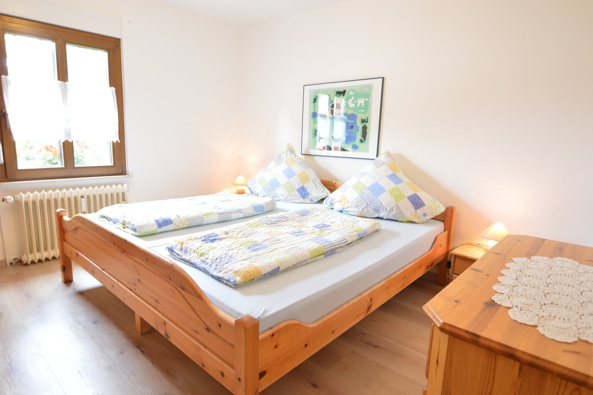Un simple lit en bois avec une couverture, une table de chevet avec une lampe, un tableau encadré au mur et une fenêtre avec des rideaux.