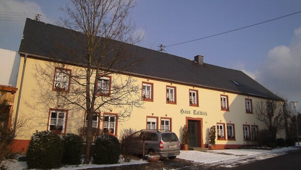 Egy kétemeletes épület, melynek sárga homlokzata és piros színű díszítőelemekkel ellátott keretei vannak, a neve „Haus Tallock”.
