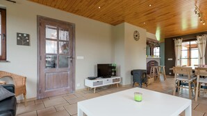 Una sala de estar con una mesa de centro blanca, un televisor sobre un mueble blanco, una puerta de madera y un comedor con sillas y una mesa de madera.