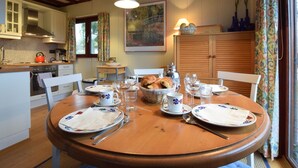 A dining area with a wooden table set for four, surrounded by chairs. There is a kitchen in the background with white cabinets and a stove.