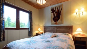 A bedroom with a large bed, wooden headboard, two bedside tables with lamps, a window with curtains, and a wall-mounted artwork.