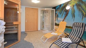 Una habitación con sauna, sillón de masaje, ducha envuelta en vidrio y un mural con temática de playa.